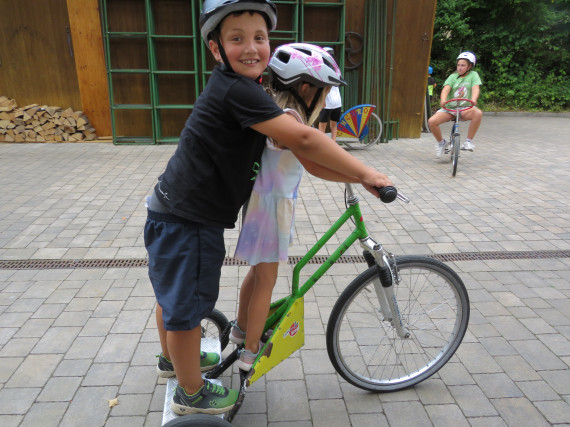 Im Vordergrund fahren zwei Kinder zusammen auf einem Rollerfahrrad, im Hintergrund ein Kind auf einem Fahrrad.